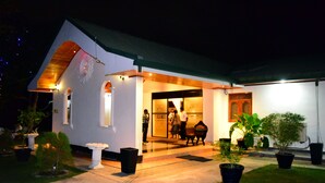 A white building with a sloped roof and a covered porch. There are potted plants and a white urn on the front lawn. - Guruge King's Island Resort (Negombo)