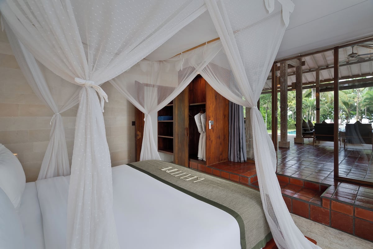 A bedroom with a canopy bed, a wooden wardrobe, and a view of a pool area.