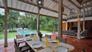Un área de comedor al aire libre techada con vista a la piscina, muebles de madera y un ventilador de techo.