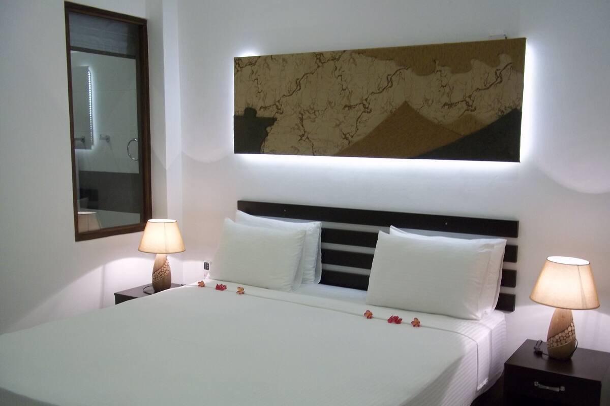 Quarto de hotel com uma cama, duas luminárias de cabeceira, uma mesa de apoio, um espelho e uma obra de arte na parede.