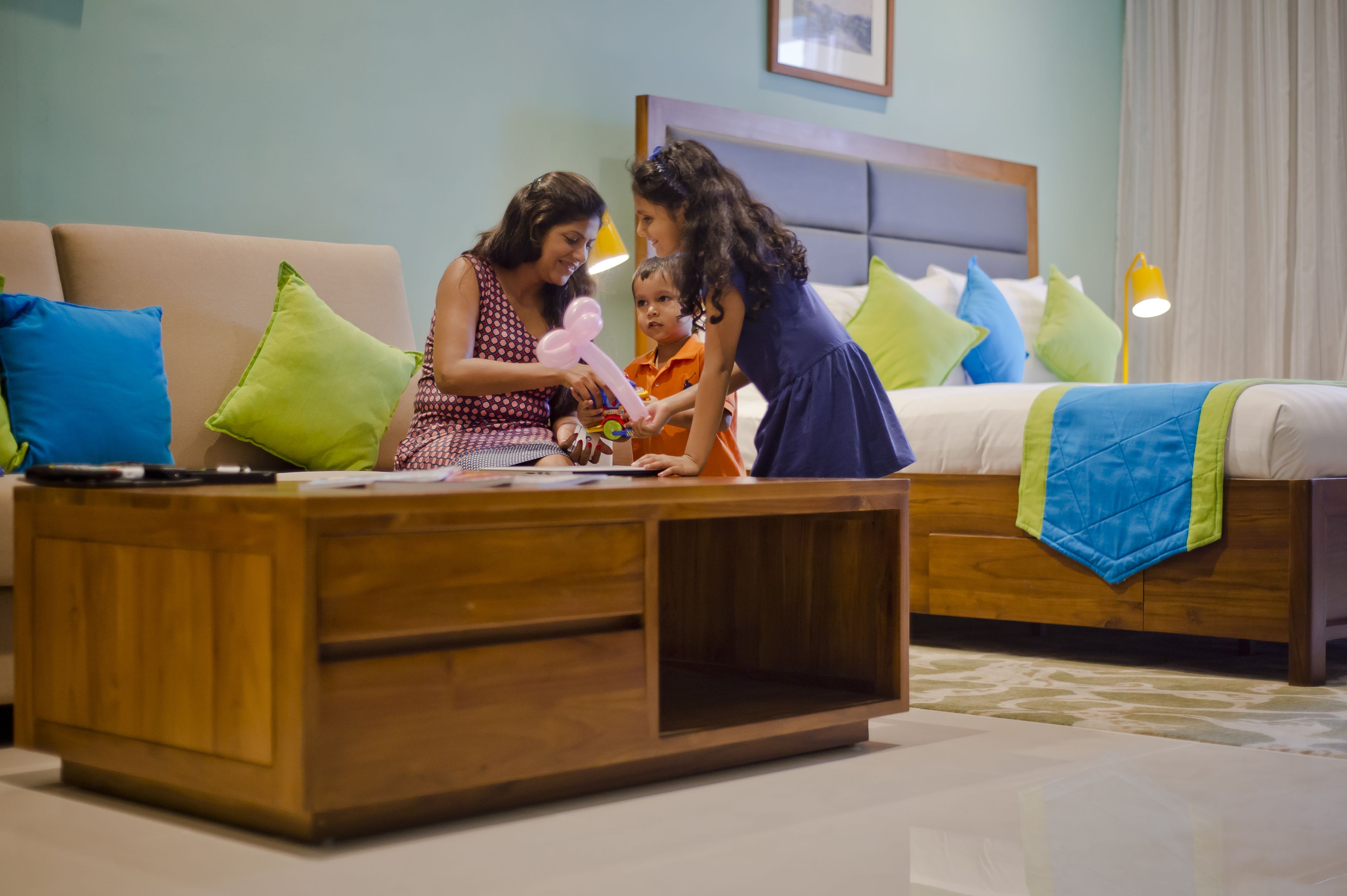 A room with a wooden coffee table, a bed with blue and green pillows, and a bedside table with a lamp.