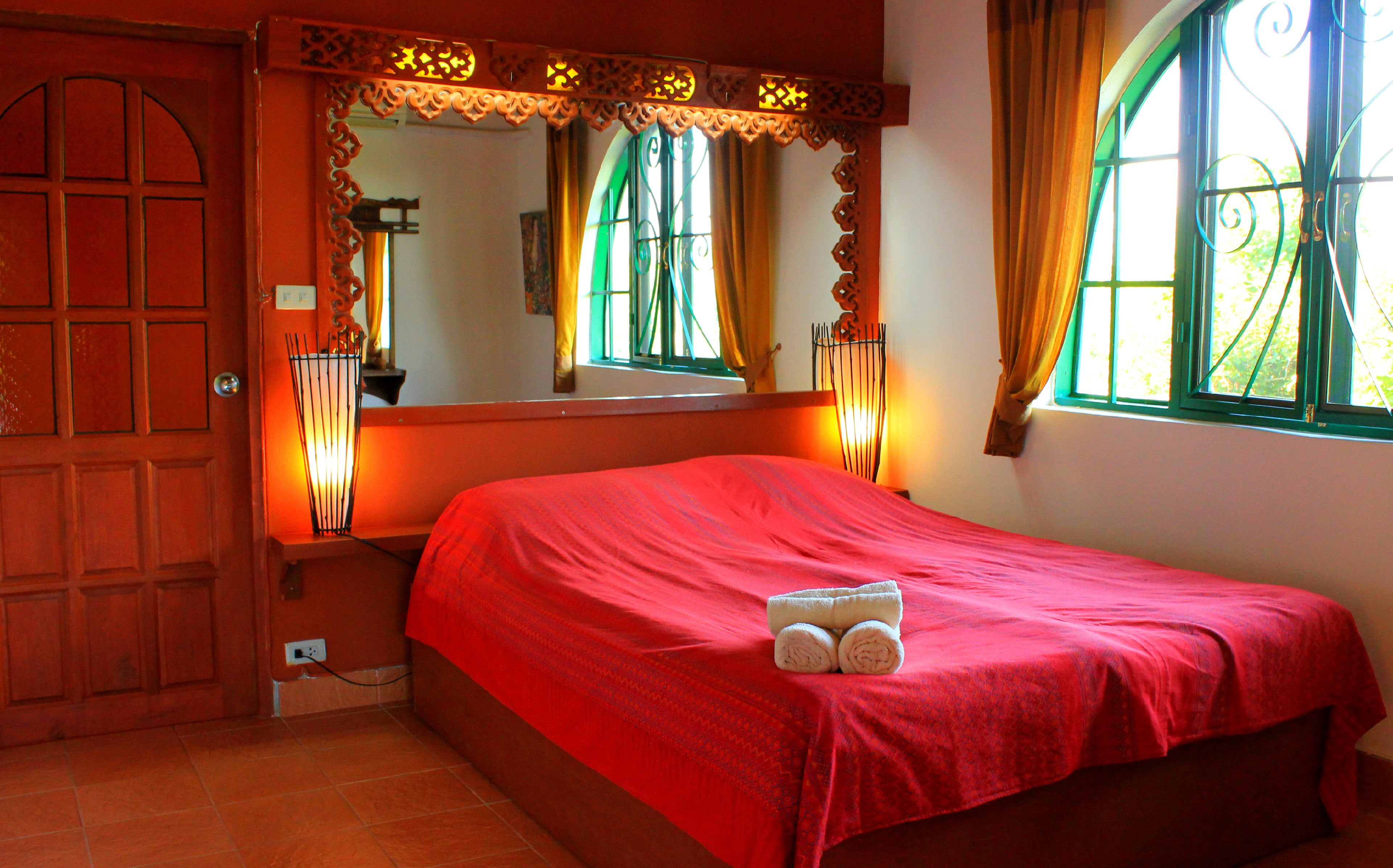 Een slaapkamer met een rode sprei, een houten deur en een raam met gordijnen.