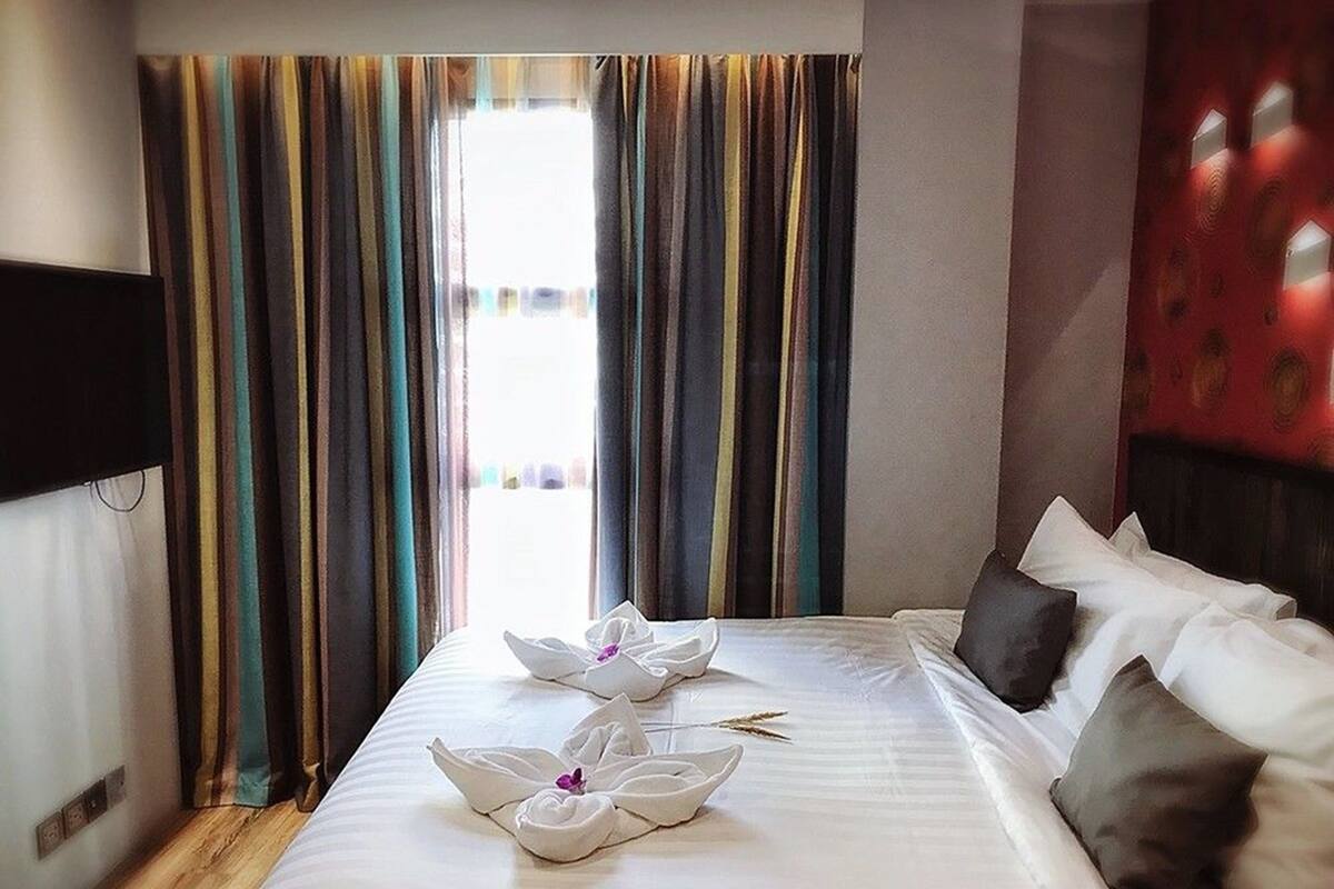 A hotel room with a neatly made bed, striped curtains, and a red wall with a geometric pattern.
