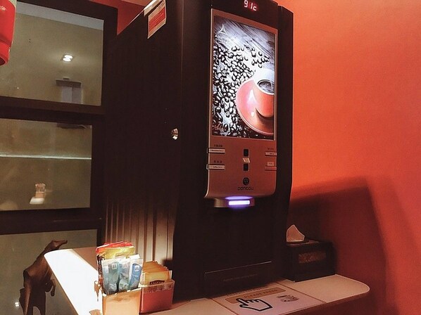 Una máquina de café con pantalla digital y botones, ubicada sobre una encimera con una caja de pañuelos y un cubo de basura cerca.