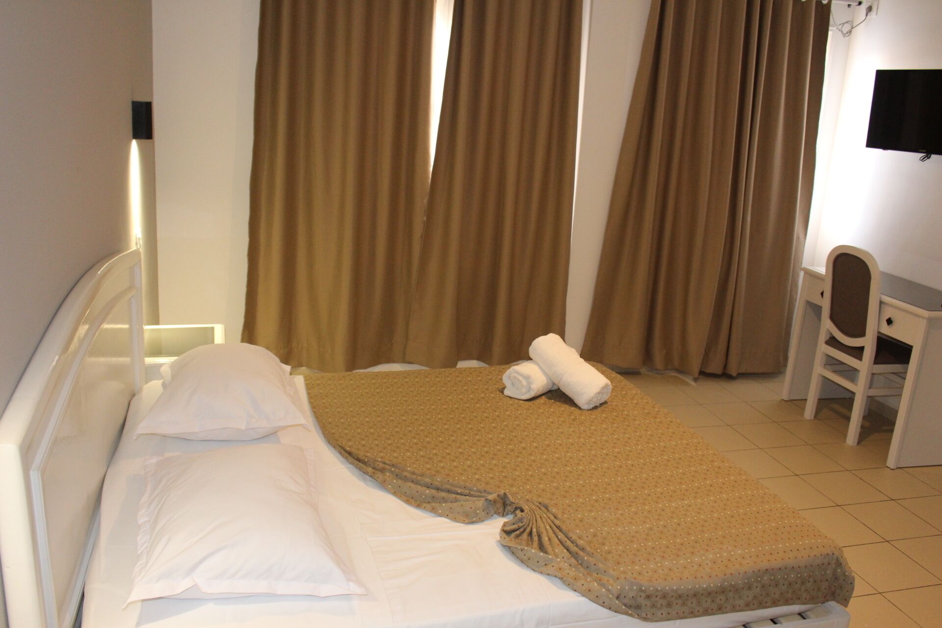 Cama bem arrumada com lençóis brancos e um cobertor marrom, uma escrivaninha pequena com uma cadeira e cortinas marrons em um quarto de hotel.