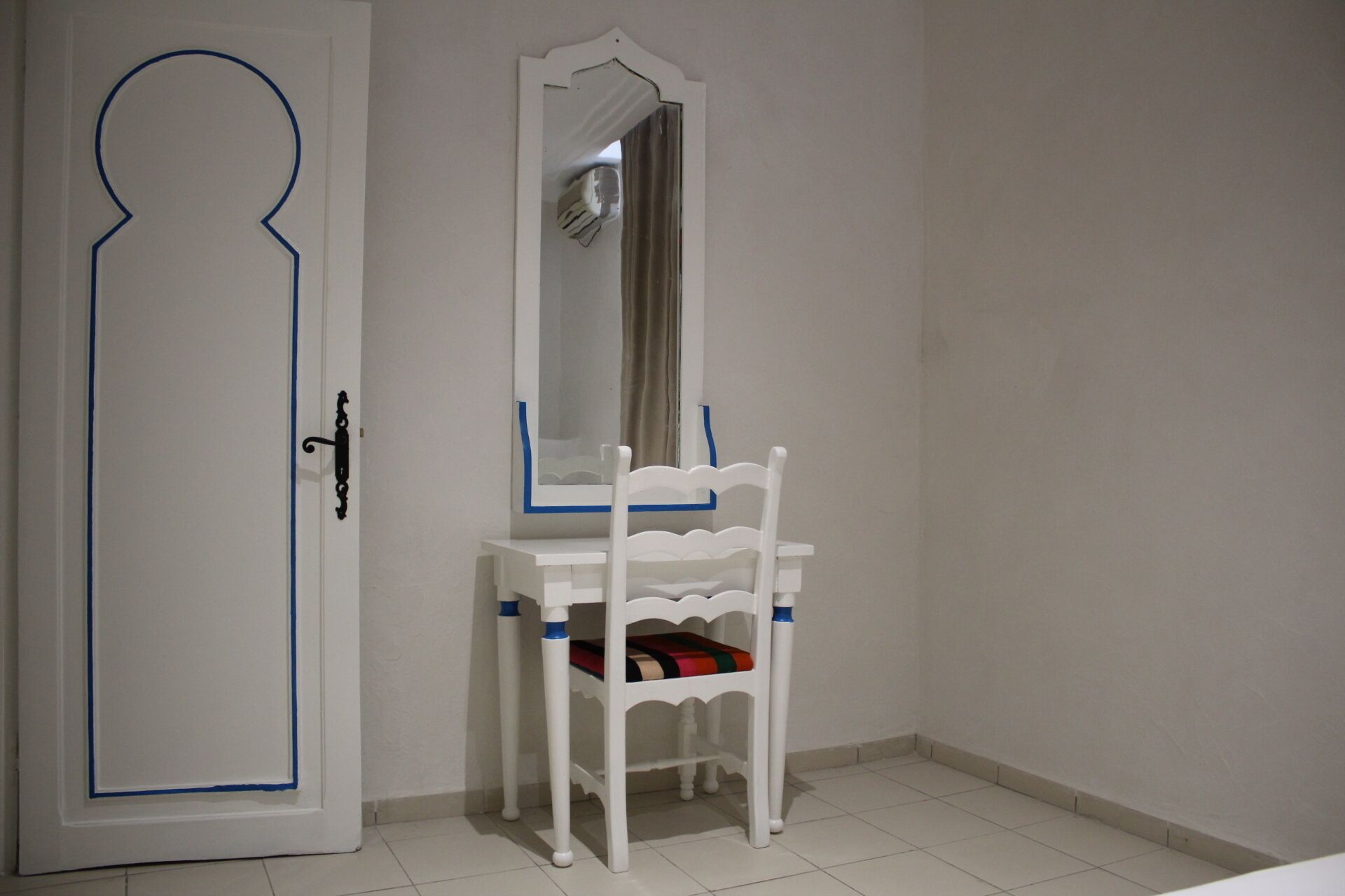 Una porta bianca con bordo blu, un comò bianco con specchio e una sedia bianca con cuscino colorato.