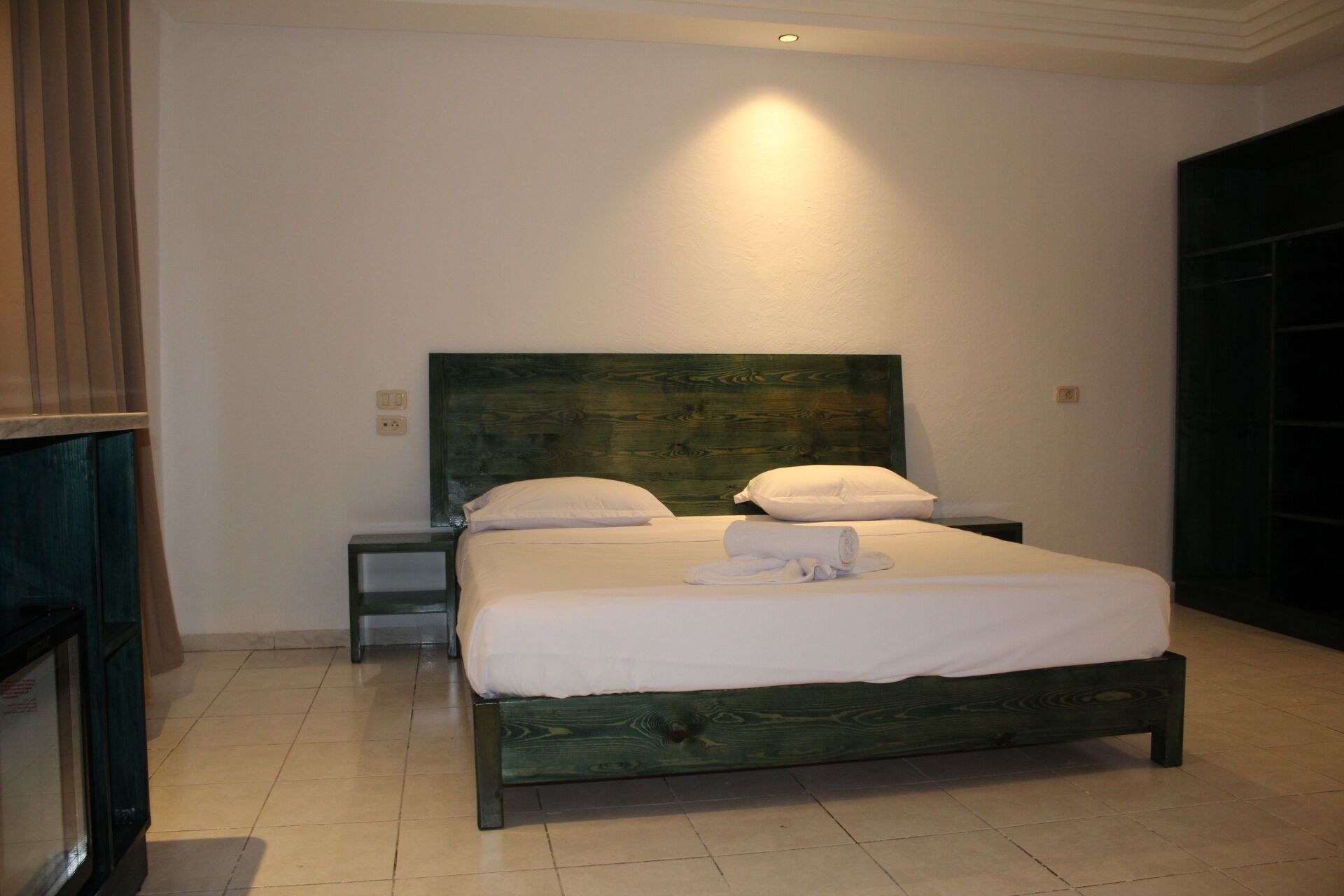 A neatly made bed with white linens and a rolled towel, a wooden headboard, a nightstand, and a dark door.
