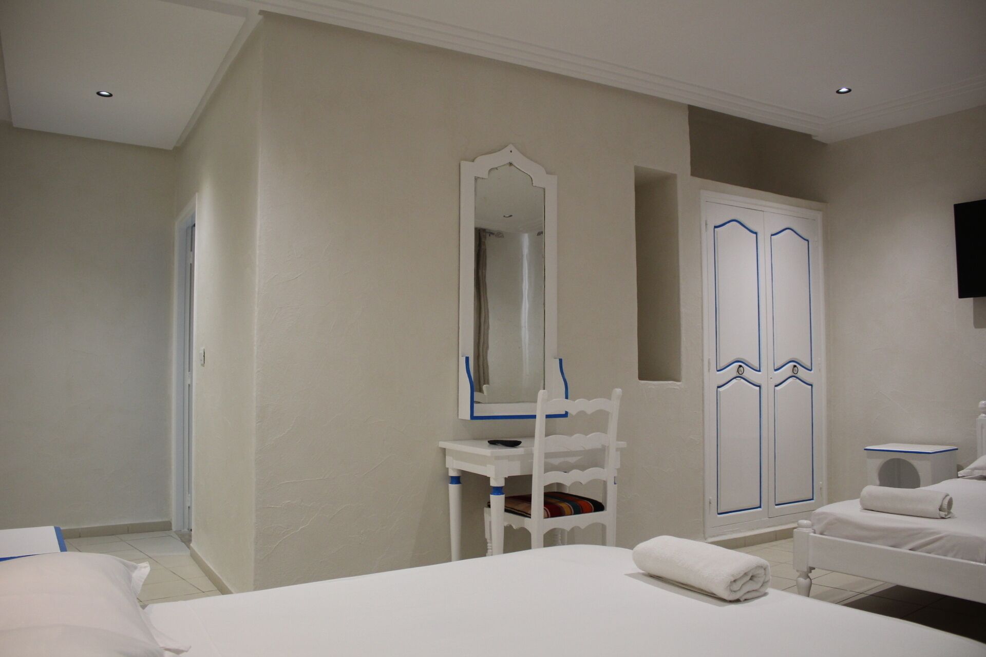 Una habitación de hotel con cama, una silla blanca, tocador con espejo y armario con detalles azules.