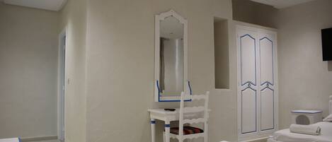 Una habitación de hotel con cama, una silla blanca, tocador con espejo y armario con detalles azules.