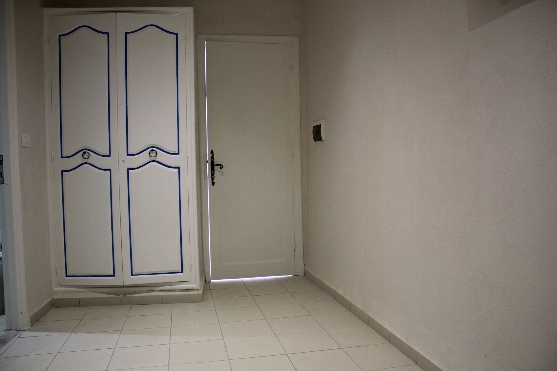 Un pasillo con una puerta blanca, detalles azules y un picaporte. El suelo está cubierto de baldosas y las paredes son lisas.