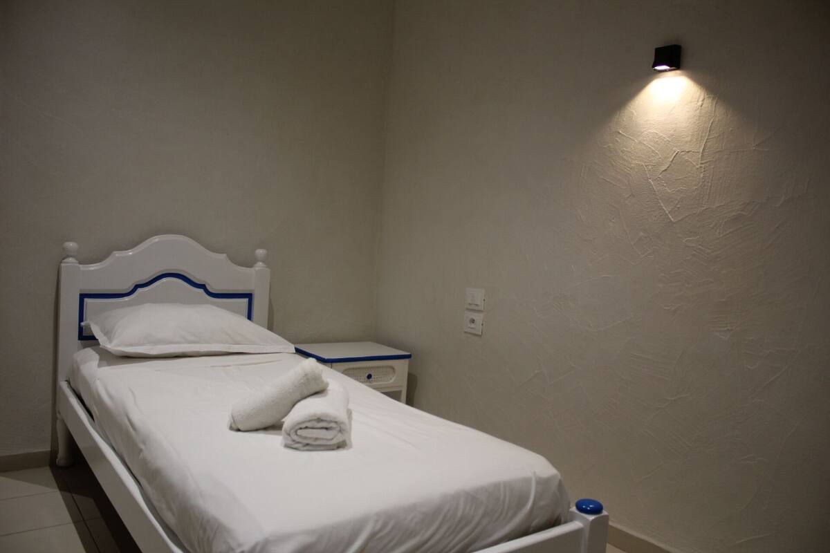 A single bed with white bedding and blue trim, a nightstand with a lamp, and a wall-mounted light fixture.