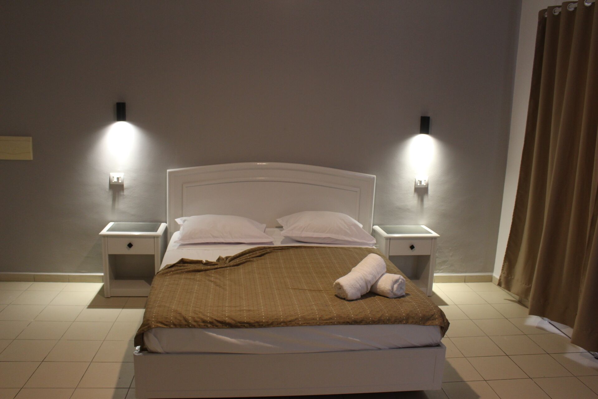 A neatly made bed with a brown blanket and white pillows, two bedside lamps, and a curtain on the right.