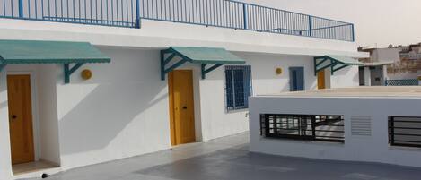 A building with a flat roof, white walls, and blue accents. There are multiple doors and windows, some with metal bars.