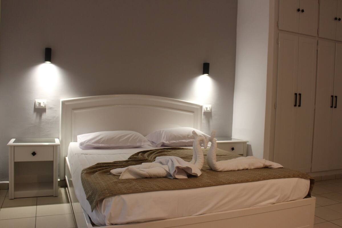 A neatly made bed with a brown blanket, two white pillows, and a white headboard. There are two bedside tables with lamps, and a wardrobe with two doors.