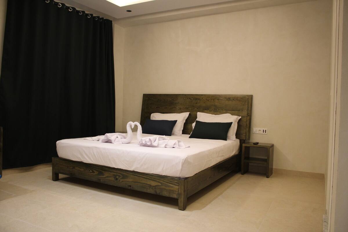 A neatly made bed with a wooden headboard, a bedside table, and a dark curtain.