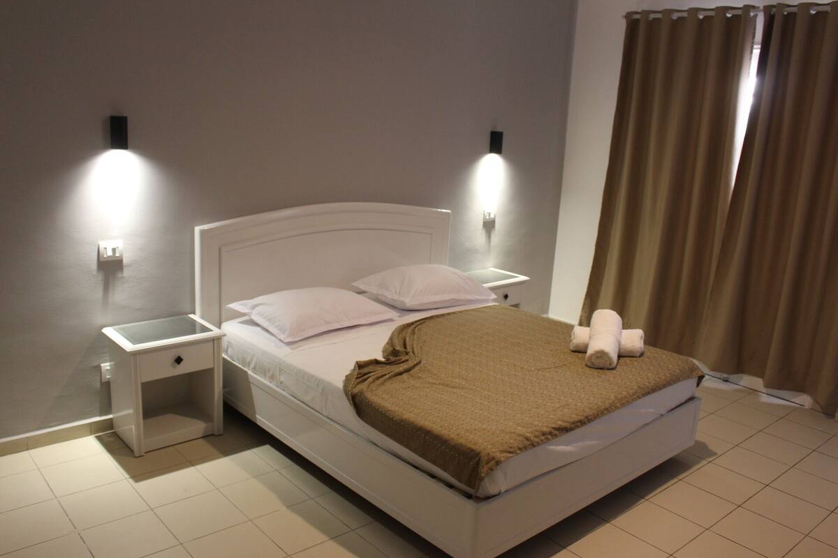 A neatly made bed with white linens and a brown throw blanket, two bedside tables with lamps, and a window with curtains.
