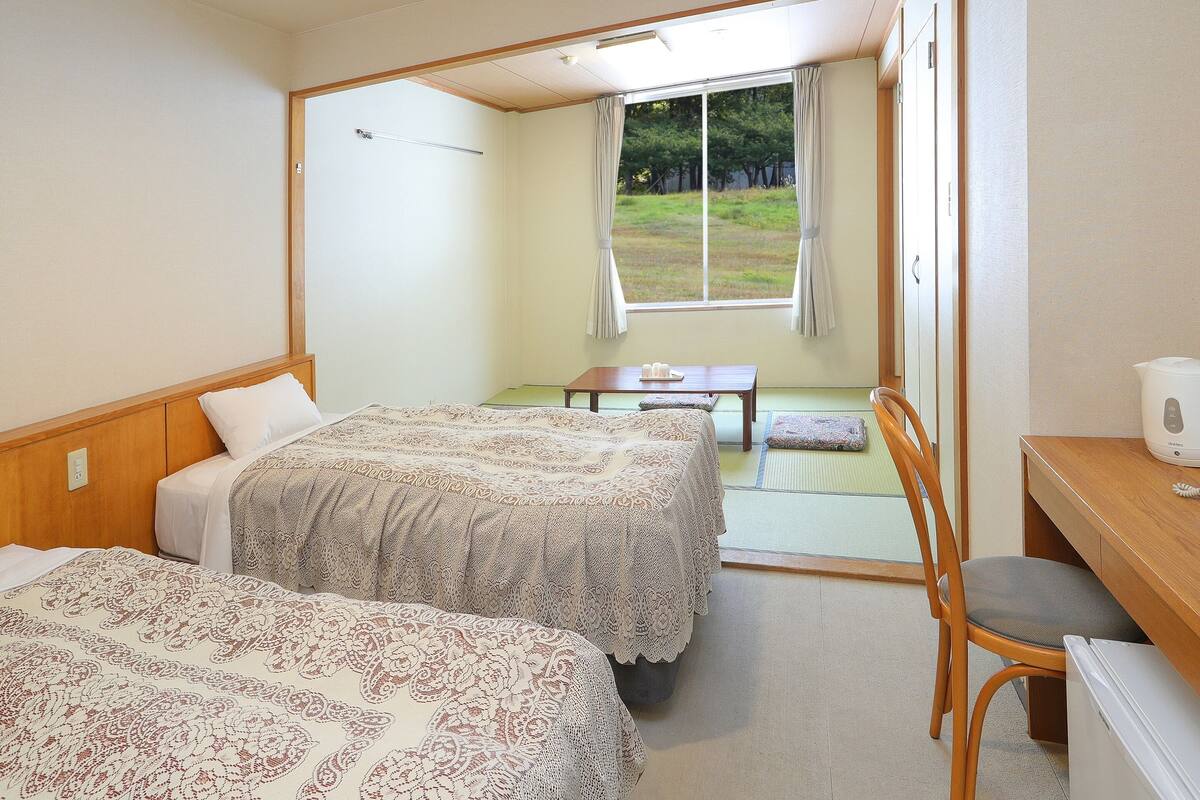 A hotel room with two beds, a small table, a chair, and a view of a grassy area through a window.