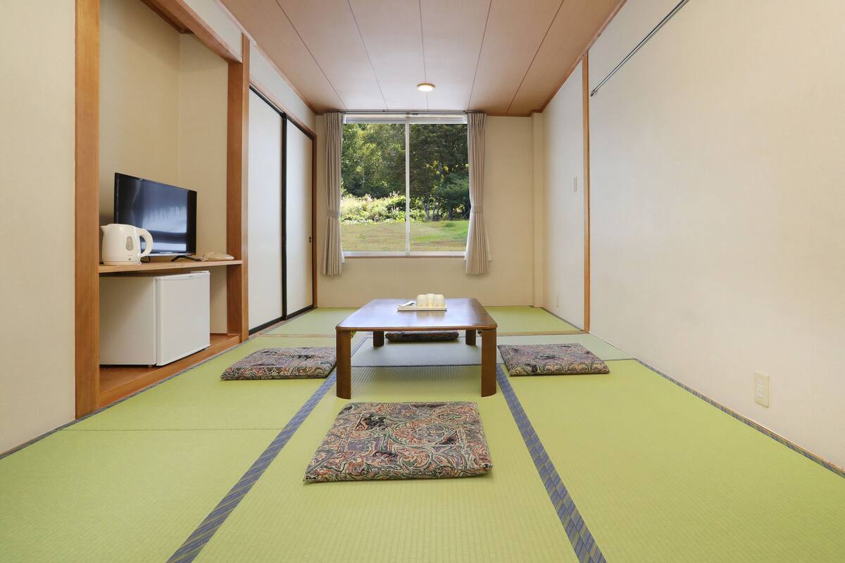 A traditional Japanese room with tatami flooring, a low table, cushions, a television, and a window with a view of greenery.
