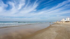 Ein weiter Sandstrand mit sanften Wellen, wolkenlosem Himmel und Gebäuden in der Ferne.
