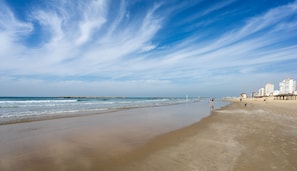 Een breed zandstrand met rustige golven, een heldere lucht en gebouwen in de verte.