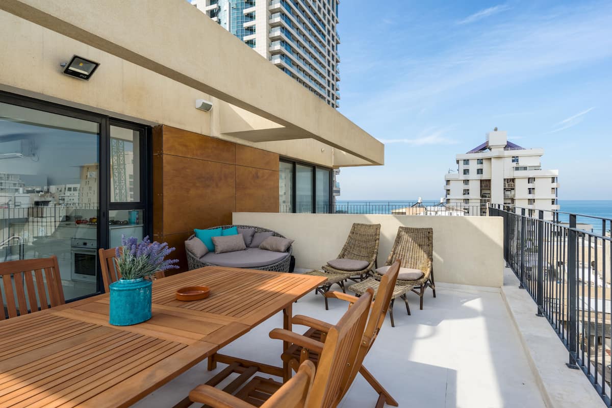 A balcony with a wooden table and chairs, a blue vase, and a view of the sea and buildings.