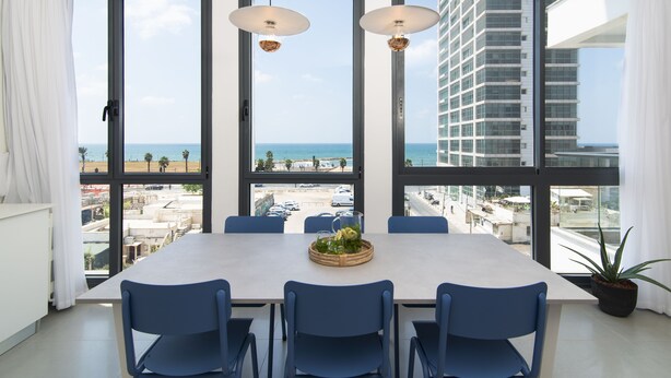 Ein Essbereich mit großem Tisch, blauen Stühlen und Blick auf das Meer und Gebäude.