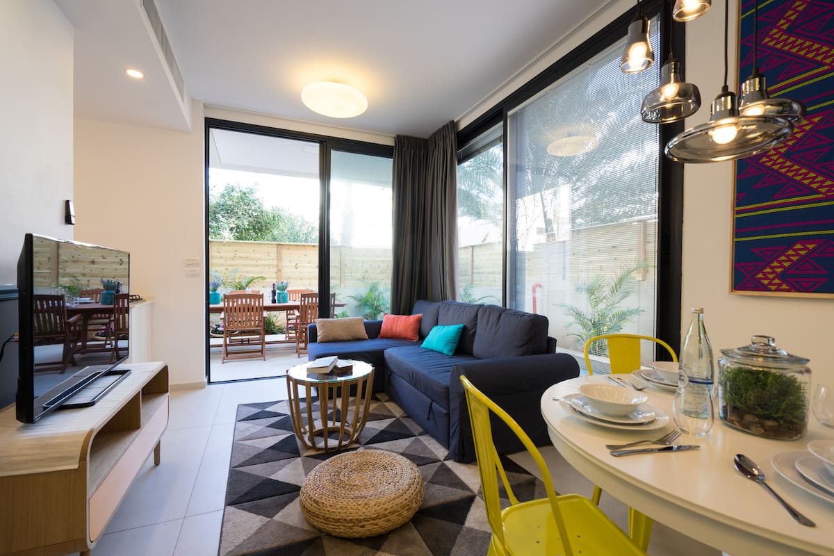 A modern living room with a sofa, dining table, and chairs, a television, and a large window with curtains.