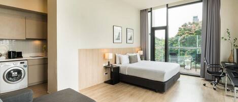 A modern hotel room with a bed, a desk, a chair, a sofa, a washing machine, and a large window with a view.