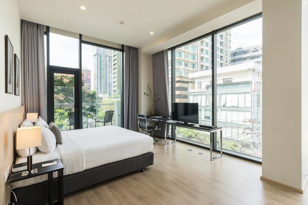 A modern hotel room with a large bed, a desk with a TV, and a balcony with a city view.