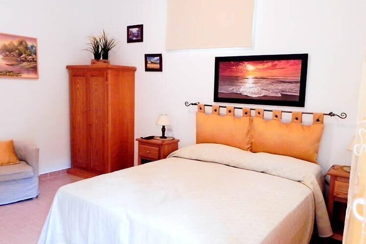 A bedroom with a bed, a nightstand, a wooden wardrobe, a chair, and a wall with framed pictures.