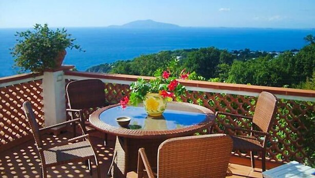 A round table with wicker chairs on a balcony overlooking a scenic view of the sea and greenery. - B&B Il Grappolo (Anacapri)