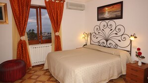 A bedroom with a bed, a nightstand, a window with curtains, a radiator, and a painting on the wall. - B&B Il Grappolo (Anacapri)