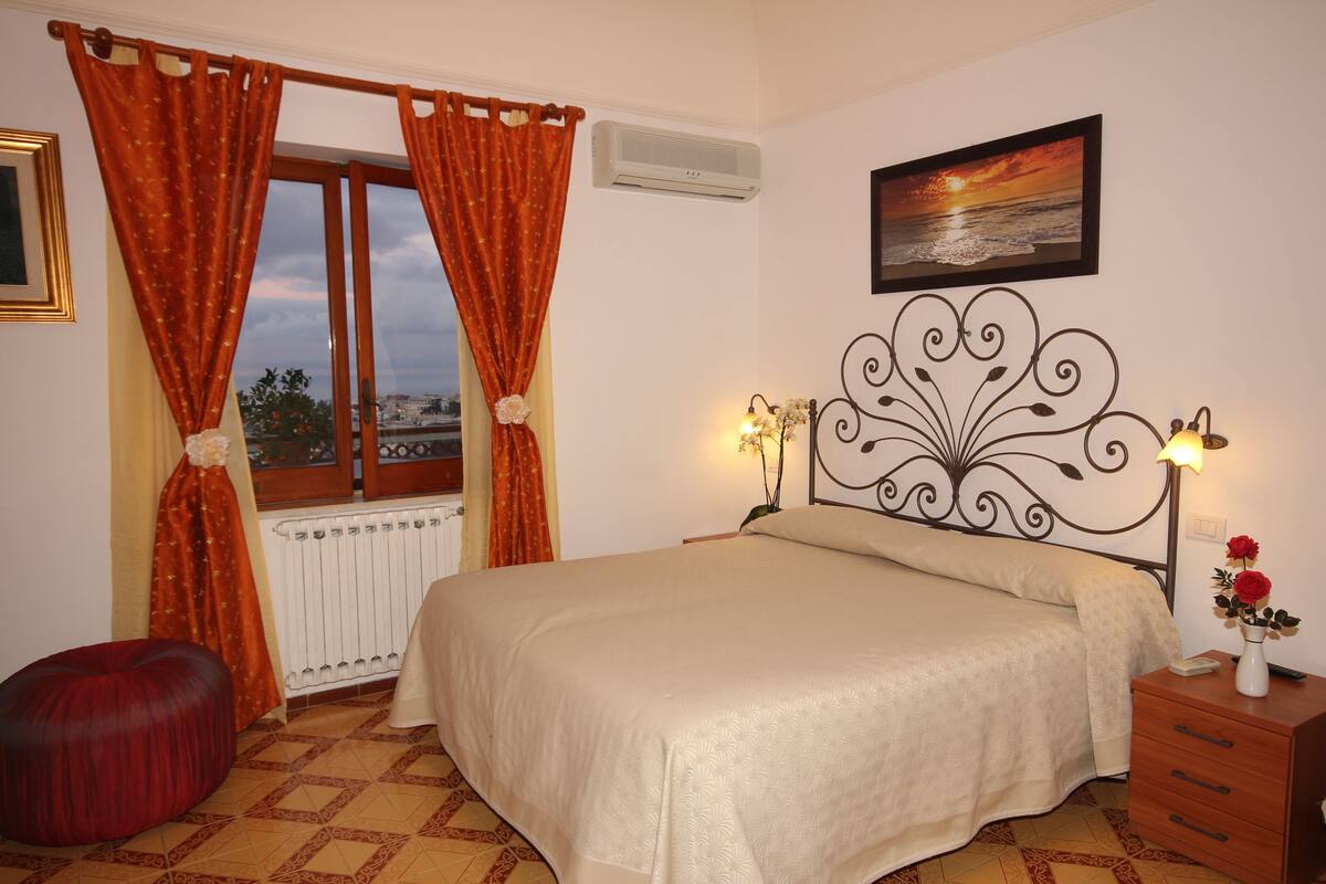 A bedroom with a bed, a nightstand, a window with curtains, a radiator, and a painting on the wall.