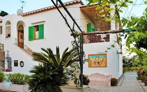 Un bâtiment à deux étages avec une façade blanche, des volets verts et un escalier menant à l’étage supérieur. On aperçoit une grande plante au premier plan et un tableau sur le mur du rez-de-chaussée.