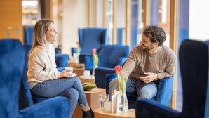 Two people seated in a modern lounge area with blue chairs and a wooden table, a vase with flowers, and a glass of water on the table. - Landhotel Weihermühle (Herschberg)