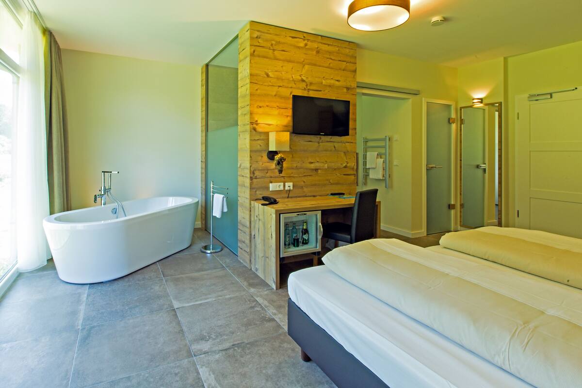 A modern hotel room with a large bathtub, a desk with a flat-screen TV, and a bed with two pillows.