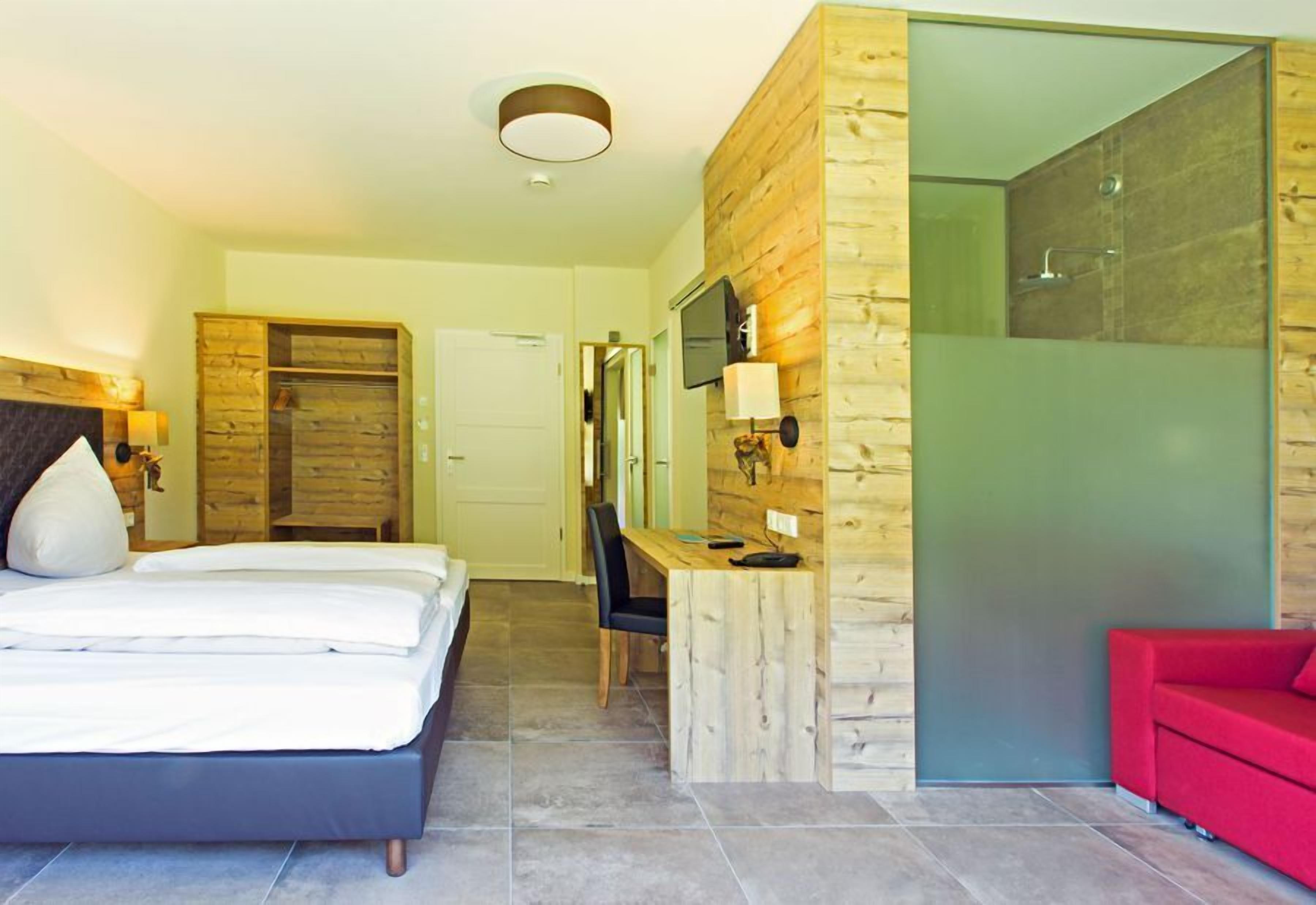 Quarto de hotel moderno com cama, escrivaninha, sofá vermelho e box de vidro.