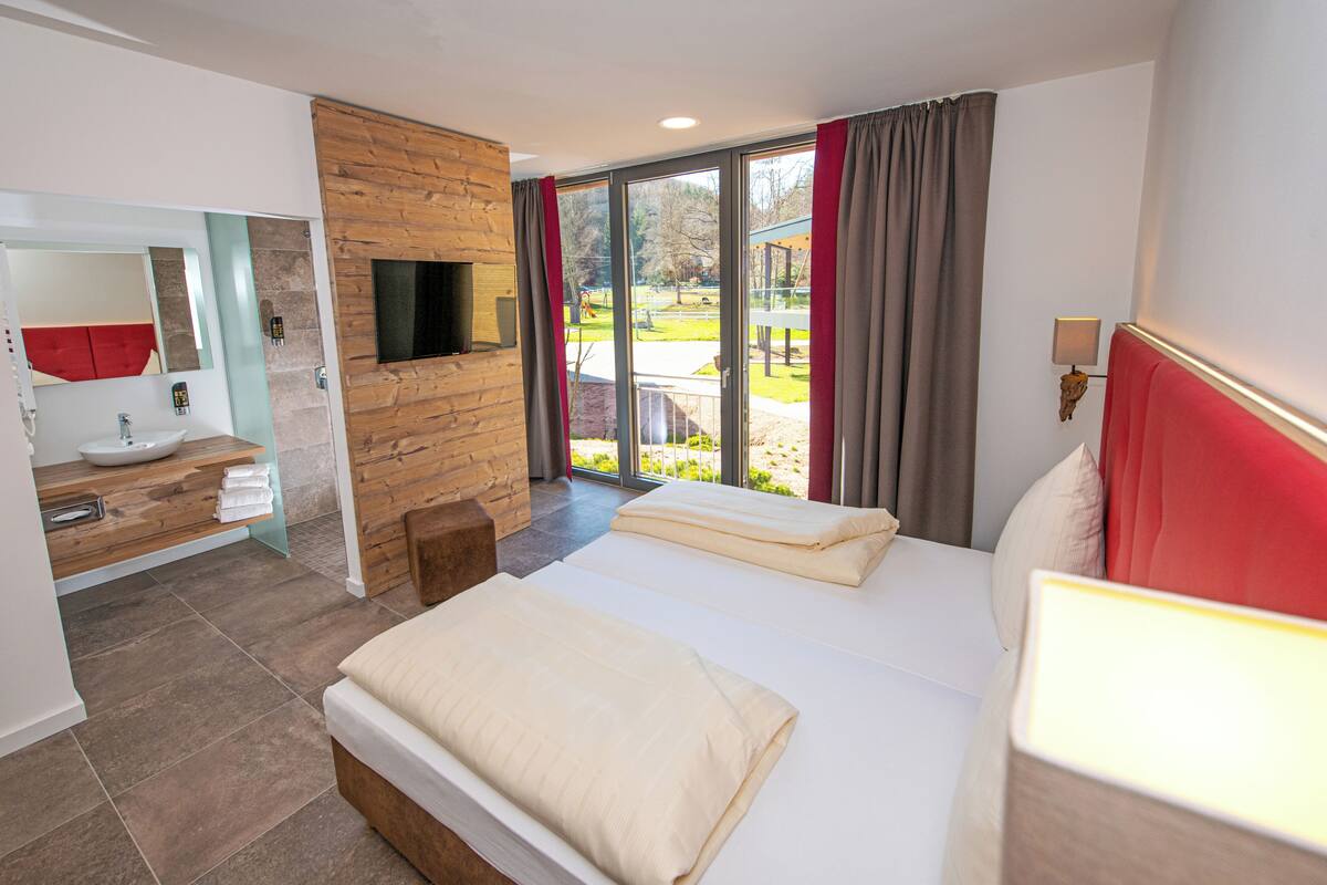 A hotel room with a bed, a TV mounted on a wooden wall, a bathroom with a sink and mirror, and a view of the outdoors.