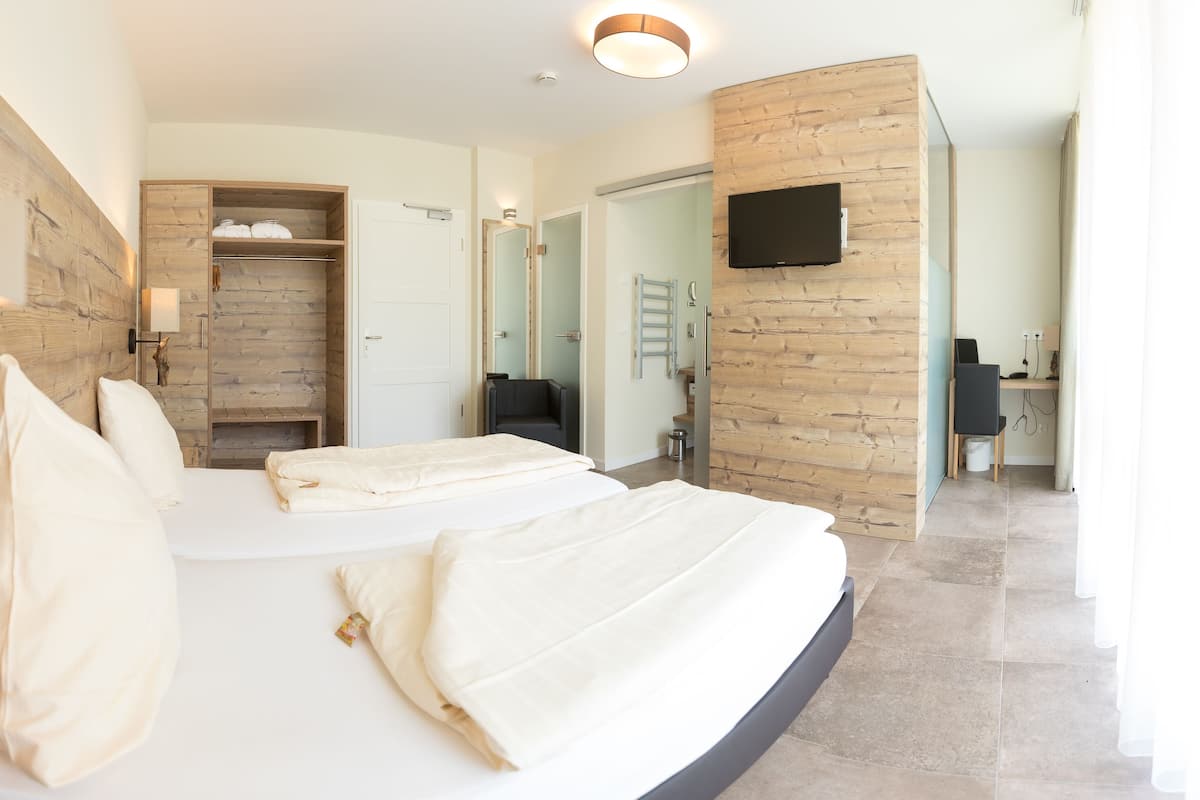 A modern hotel room with a curved bed, a wooden wardrobe, a television mounted on the wall, and a bathroom visible through an open door.