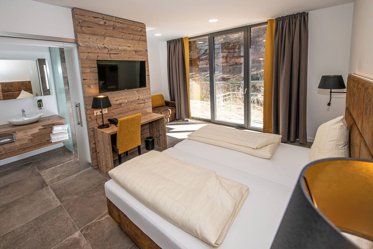 A modern hotel room with a large bed, a wooden desk, a bathroom with a sink and mirror, and a TV mounted on the wall.