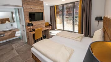 Una habitación de hotel moderna con una cama grande, un escritorio de madera, un baño con lavamanos y espejo, y un televisor montado en la pared.