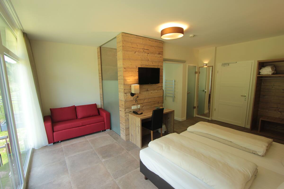 A modern hotel room with a large bed, a red sofa, a desk with a chair, a TV mounted on a wooden wall, and a bathroom visible through an open door.