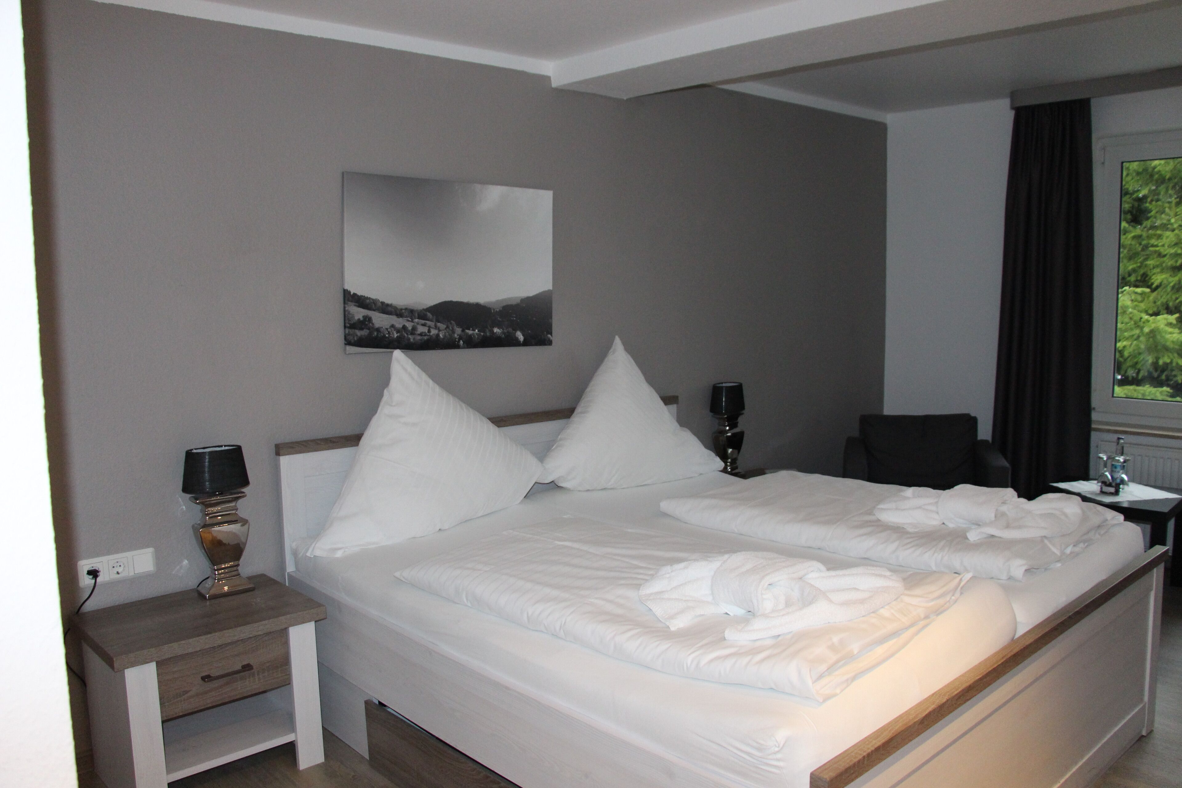 A hotel room with a large bed, a bedside table with a lamp, a chair, a small table with a vase, and a window with curtains.