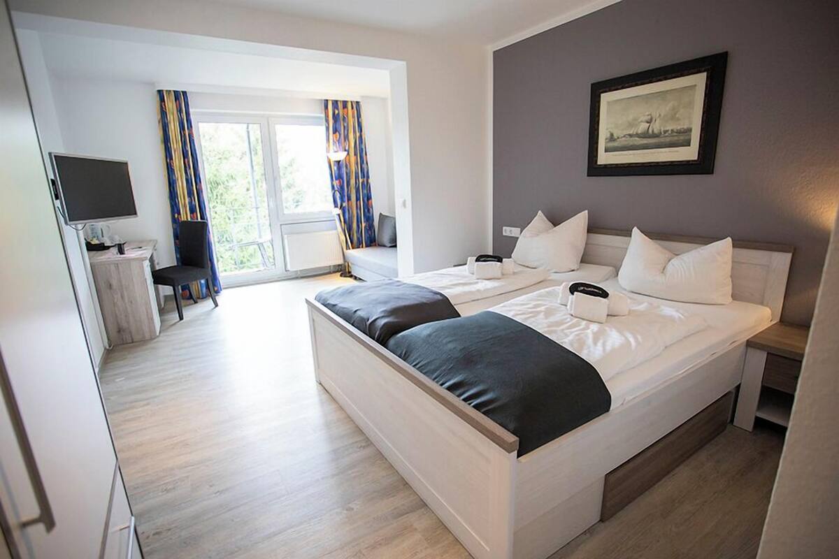 A hotel room with a bed, a desk with a computer, a chair, and a window with curtains.