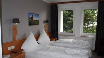 A hotel room with a large bed, two bedside tables, a nightstand, a window with a view of trees, and two wall-mounted lamps.