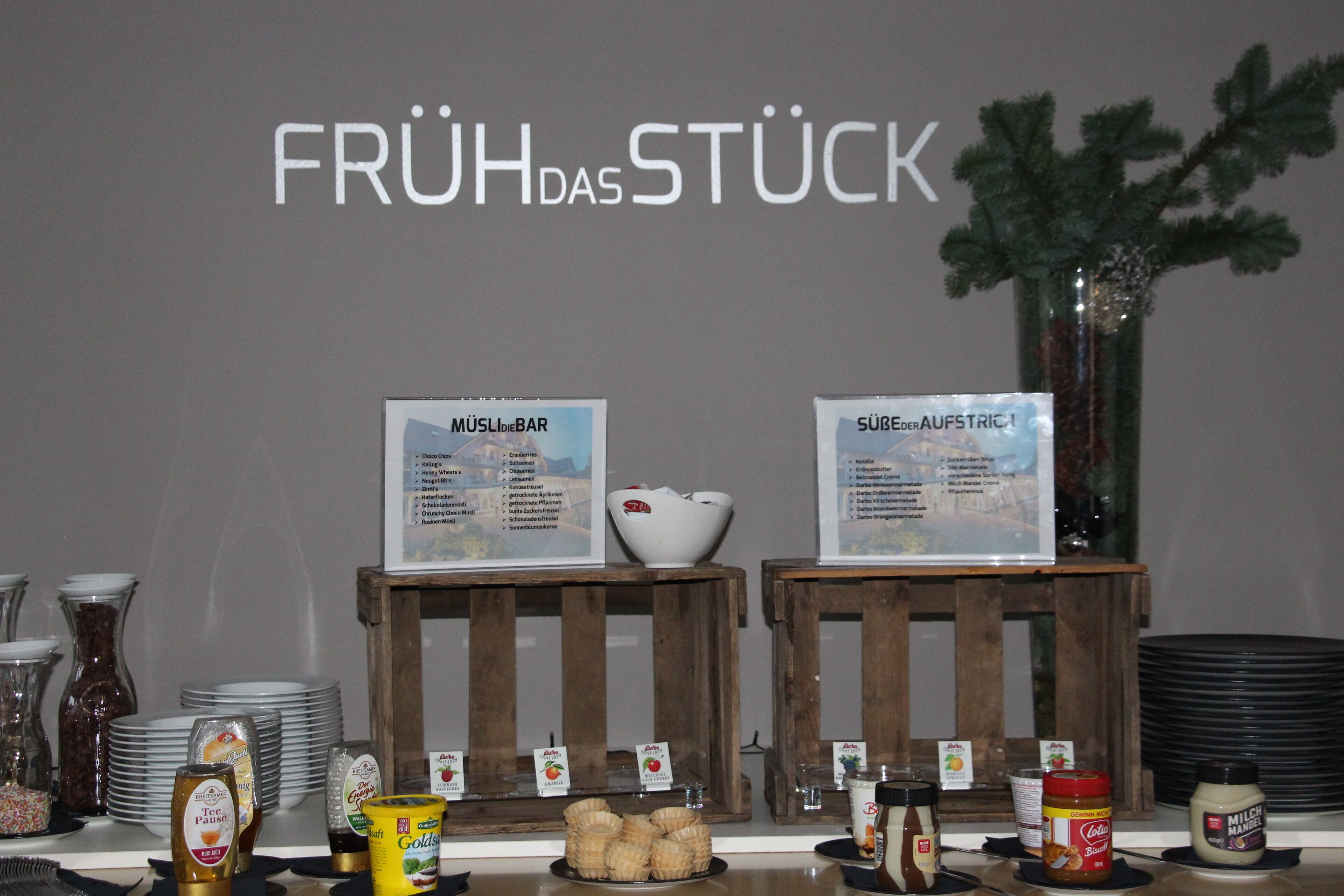 Een ontbijtbuffet met daarin verschillende gerechten met labels, en een bordje met de tekst 'FRÜH DAS STÜCK'.