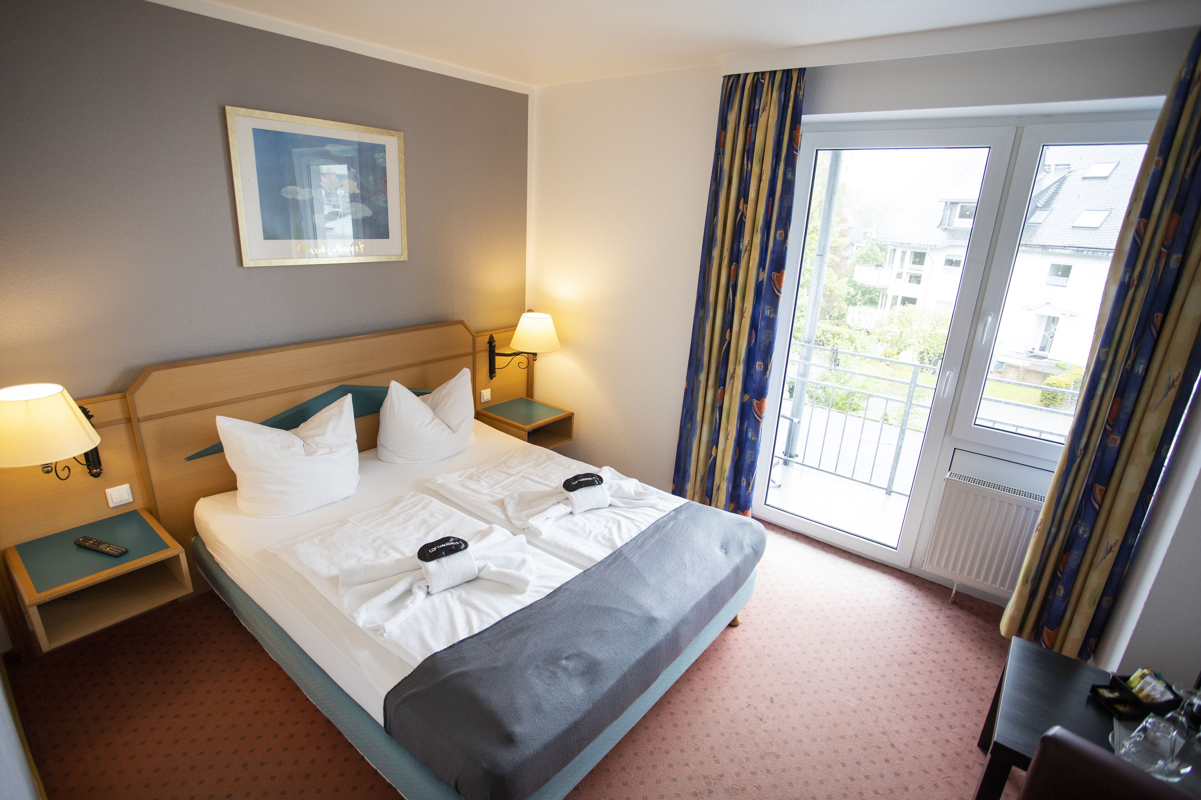 Hotelkamer met een bed, twee nachtkastjes, een lamp, een inlijsting foto aan de muur en uitzicht buiten door een raam met gordijnen.