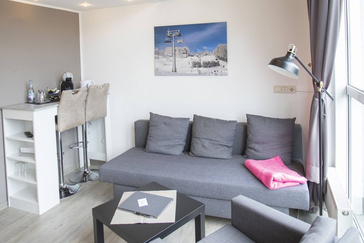 A modern living room with a grey sofa, a small black table, a staircase with wooden steps, and a window with curtains.