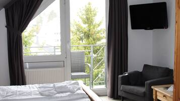 Un dormitorio con una cama de madera, un sofá gris, un televisor en la pared y un balcón con vistas a árboles.