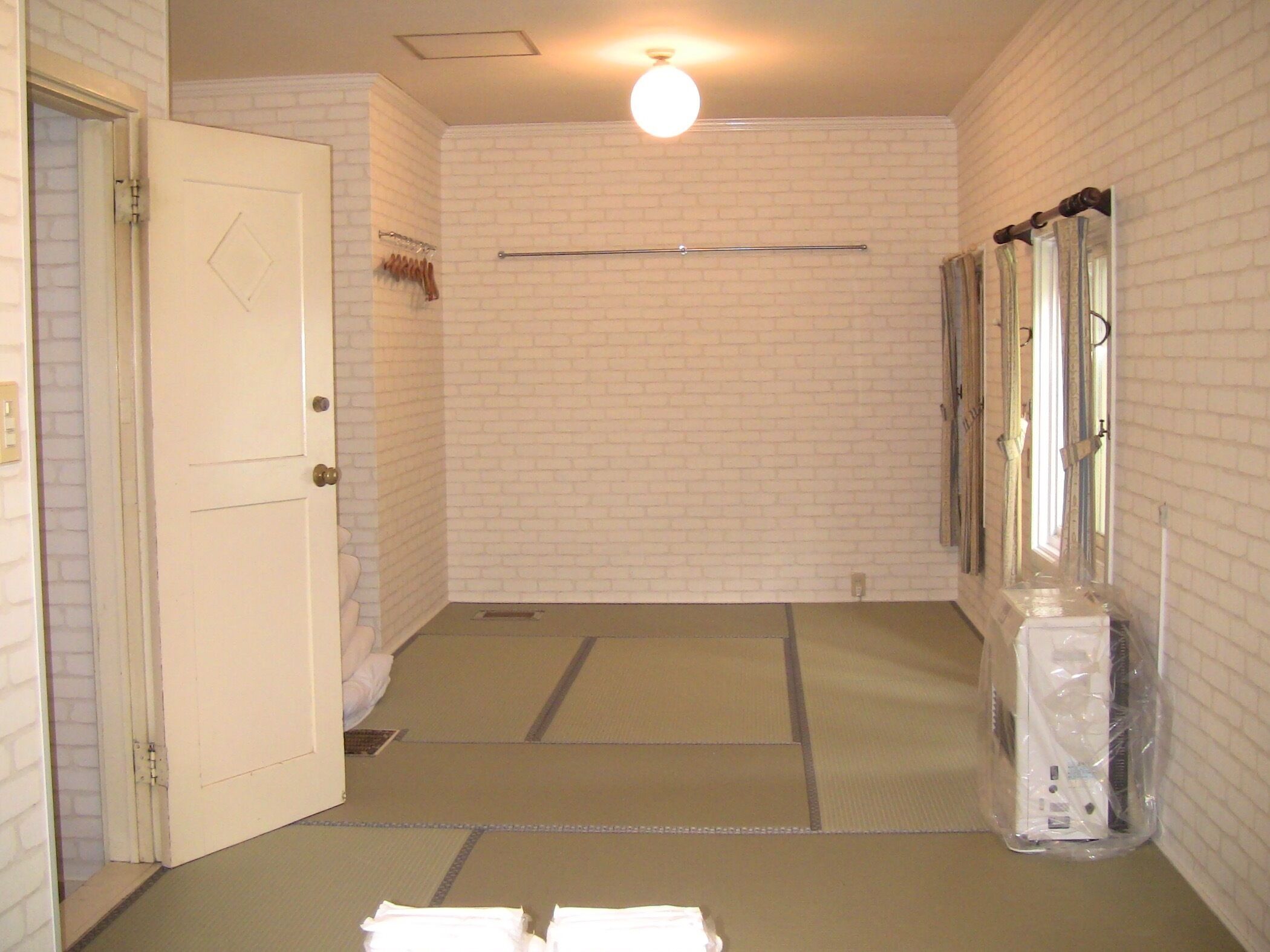 Quarto de hotel com parede de tijolos brancos, porta, porta-toalhas, toalha no chão e janela com cortinas.
